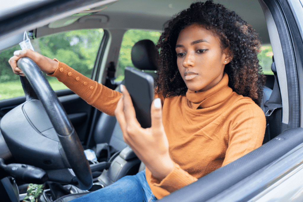 A black woman sitting in a car holding the steering while looking at her phone