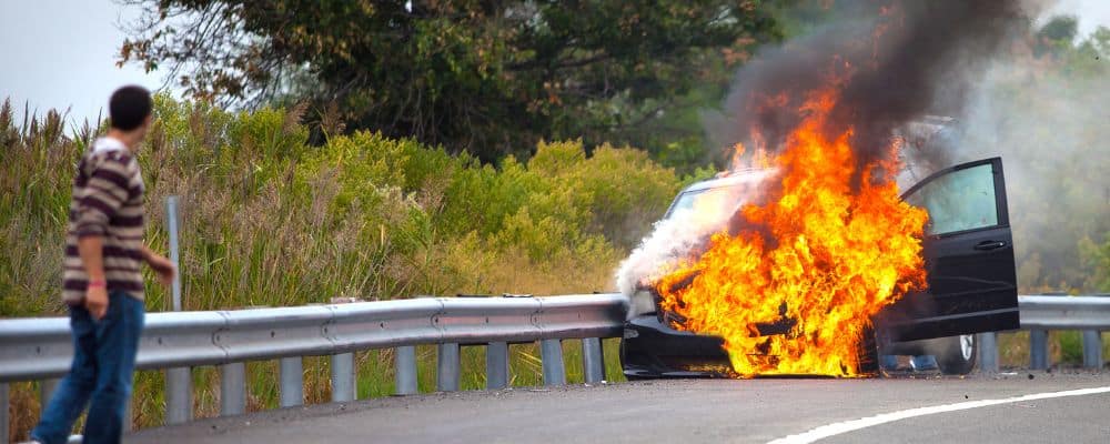 A person approaches a car on fire on the side of the road
