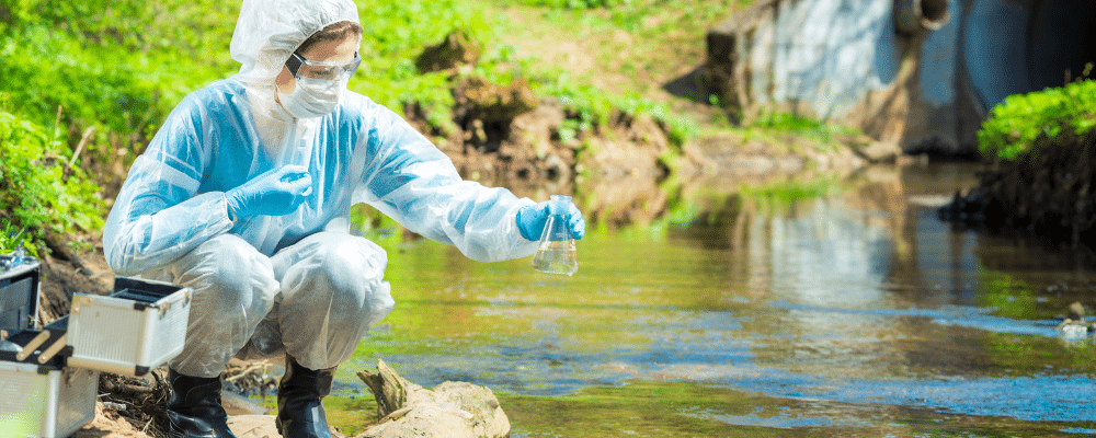 a person in a protective suit and boots, sitting on the bank of a river and testing the water for contaminants